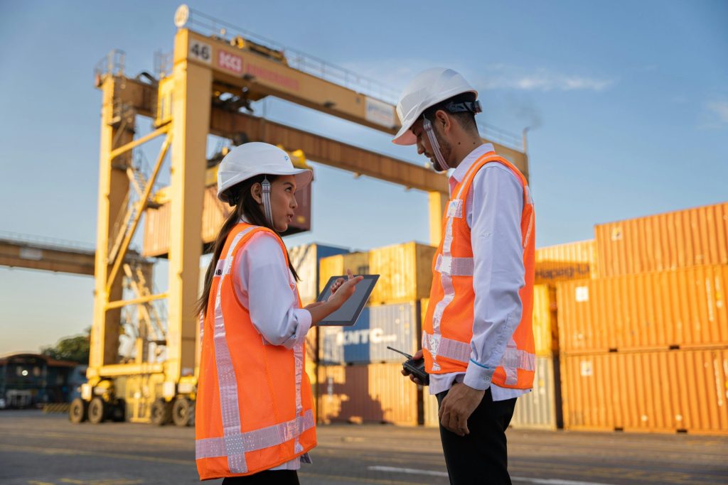 Two workers in hard hats discuss plans near shipping containers